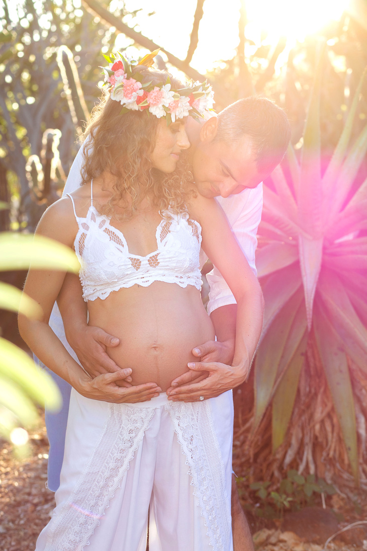 Headshots, Maternity, Child & Family Photography -  Oahu, Hawaii - happy couple with husband holding pregnant wife's belly with sunshine behind them in a garden